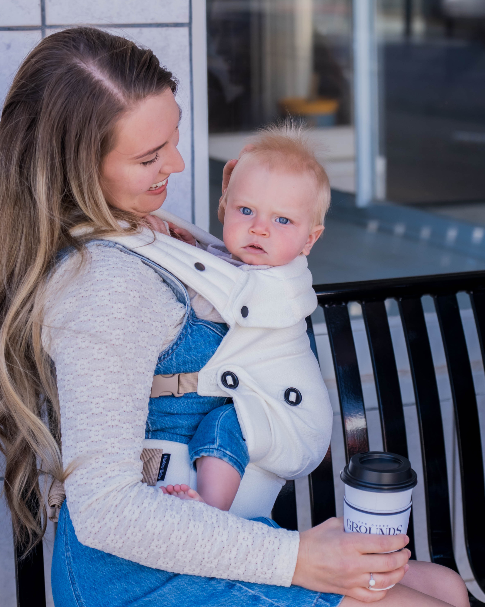 Woman holding a baby in a carrier and a coffee cup outdoors.