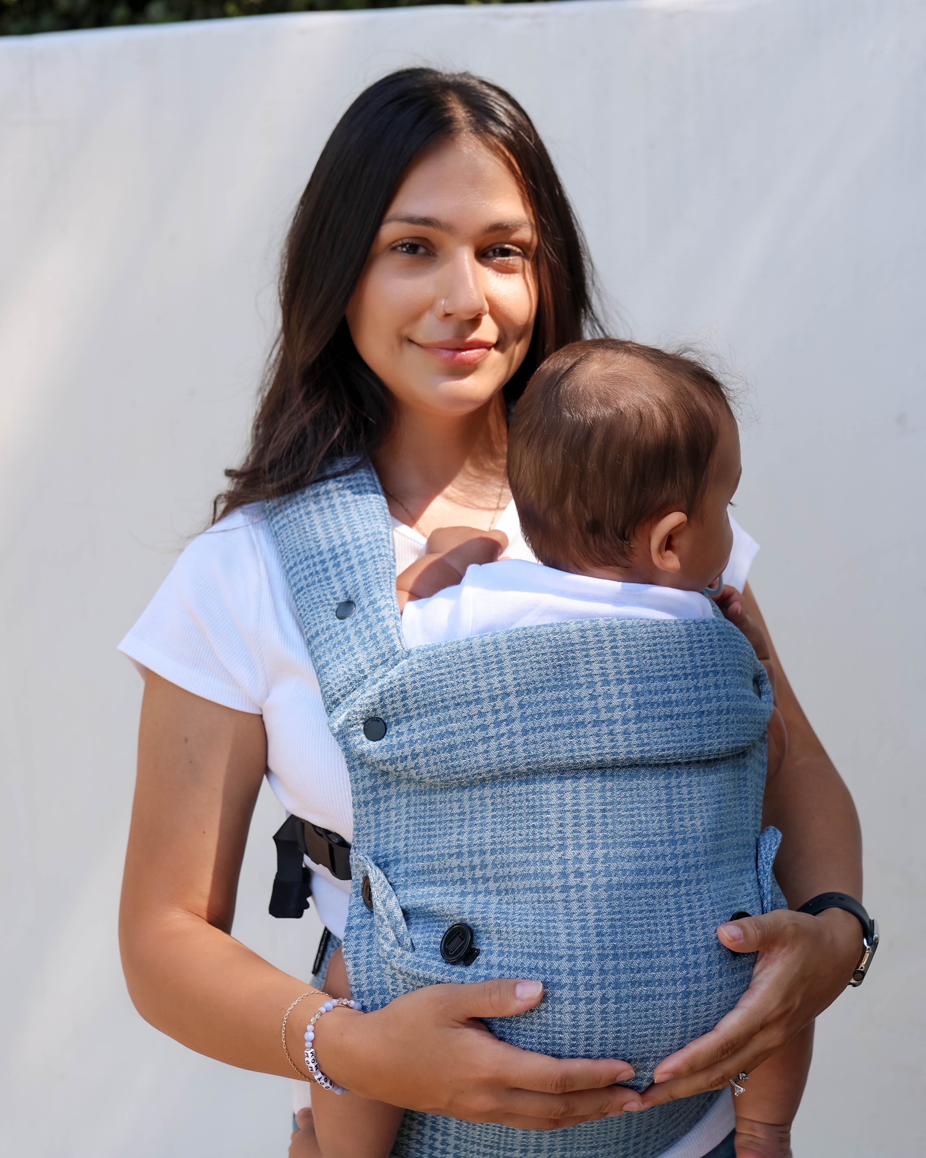 A woman wearing a white t-shirt carries a baby in a blue and white plaid wrap-style baby carrier vest against a plain white background.
