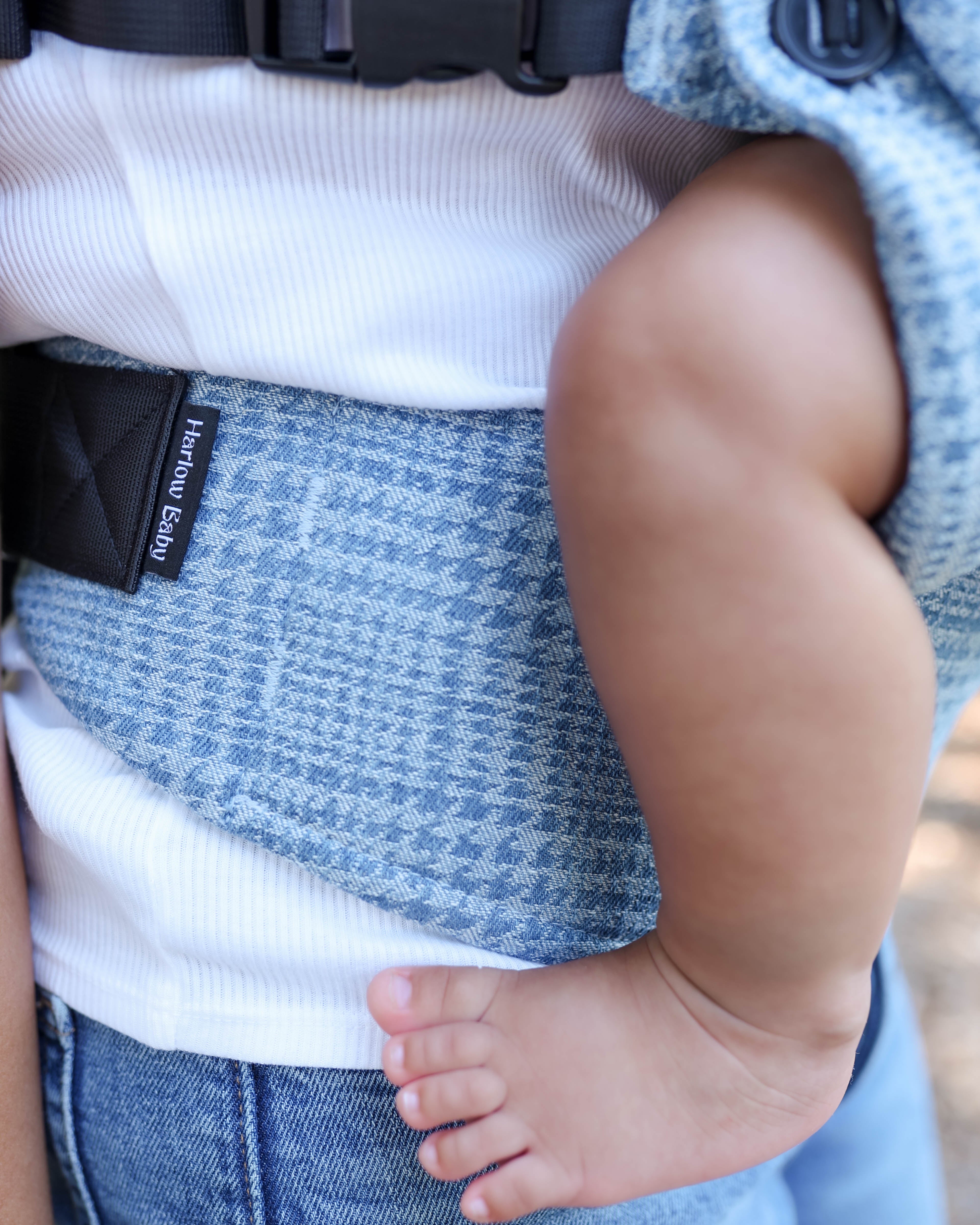 Close-up of a baby carrier worn on a person's torso, with a child's arm and foot visible. The carrier has a blue and white textured pattern, a black strap, and a small black label reading 'Harlow Baby'