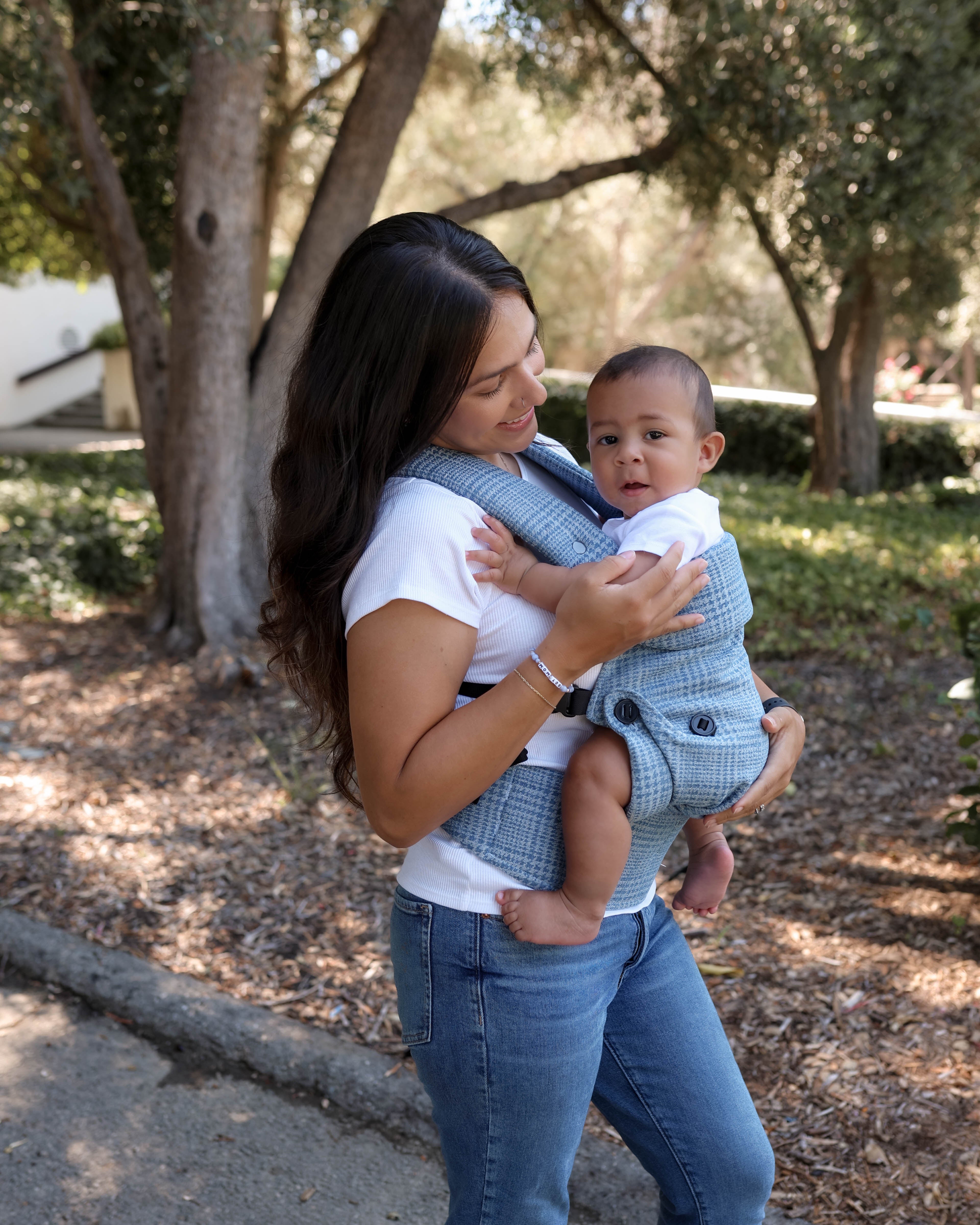 A woman outdoors carrying a baby in a blue and white checkered baby carrier wrap, set against a background of trees and a paved path.