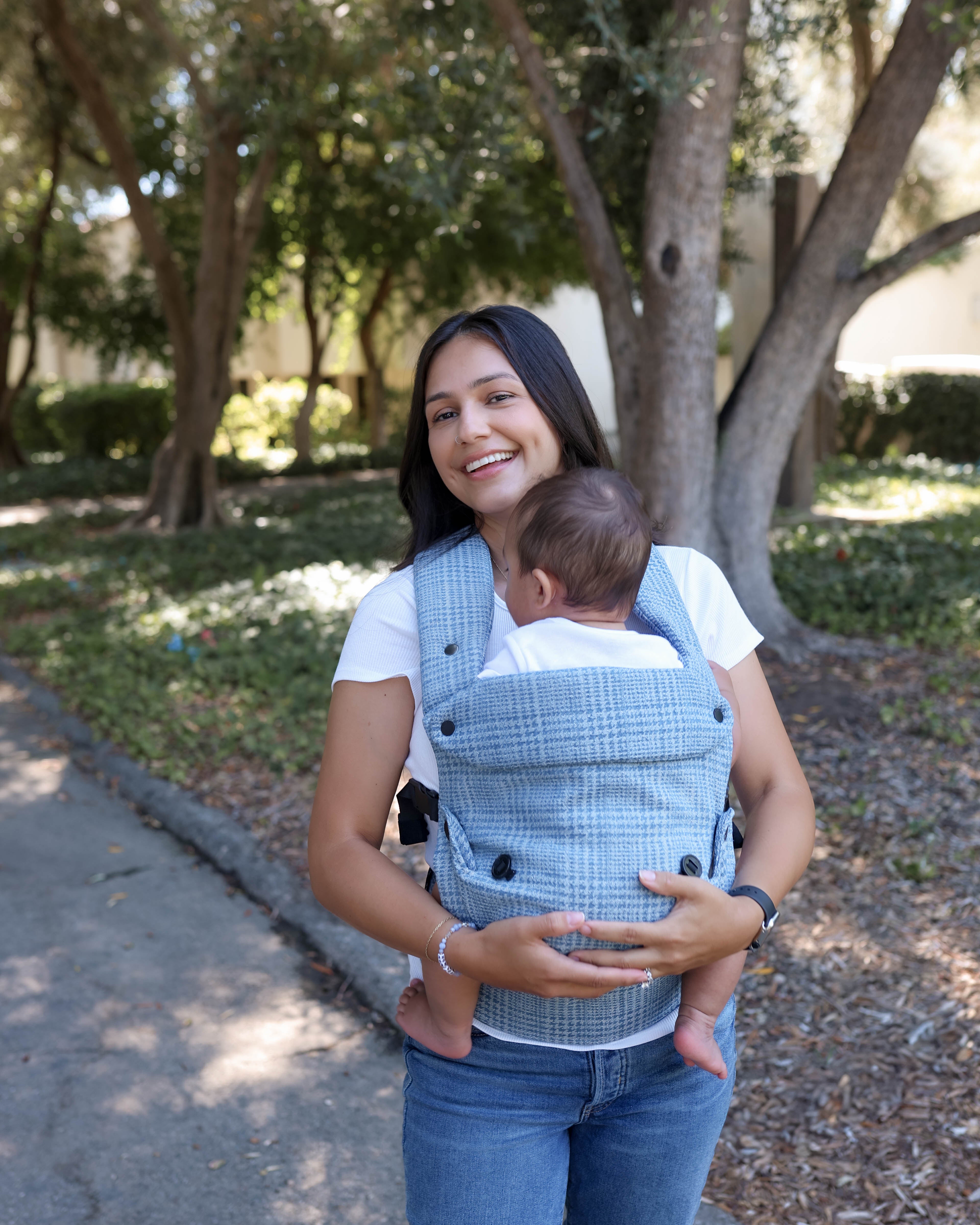 A woman wearing a light blue and white jacquard denim baby carrier with a baby inside, standing outdoors on a paved path with trees in the background.