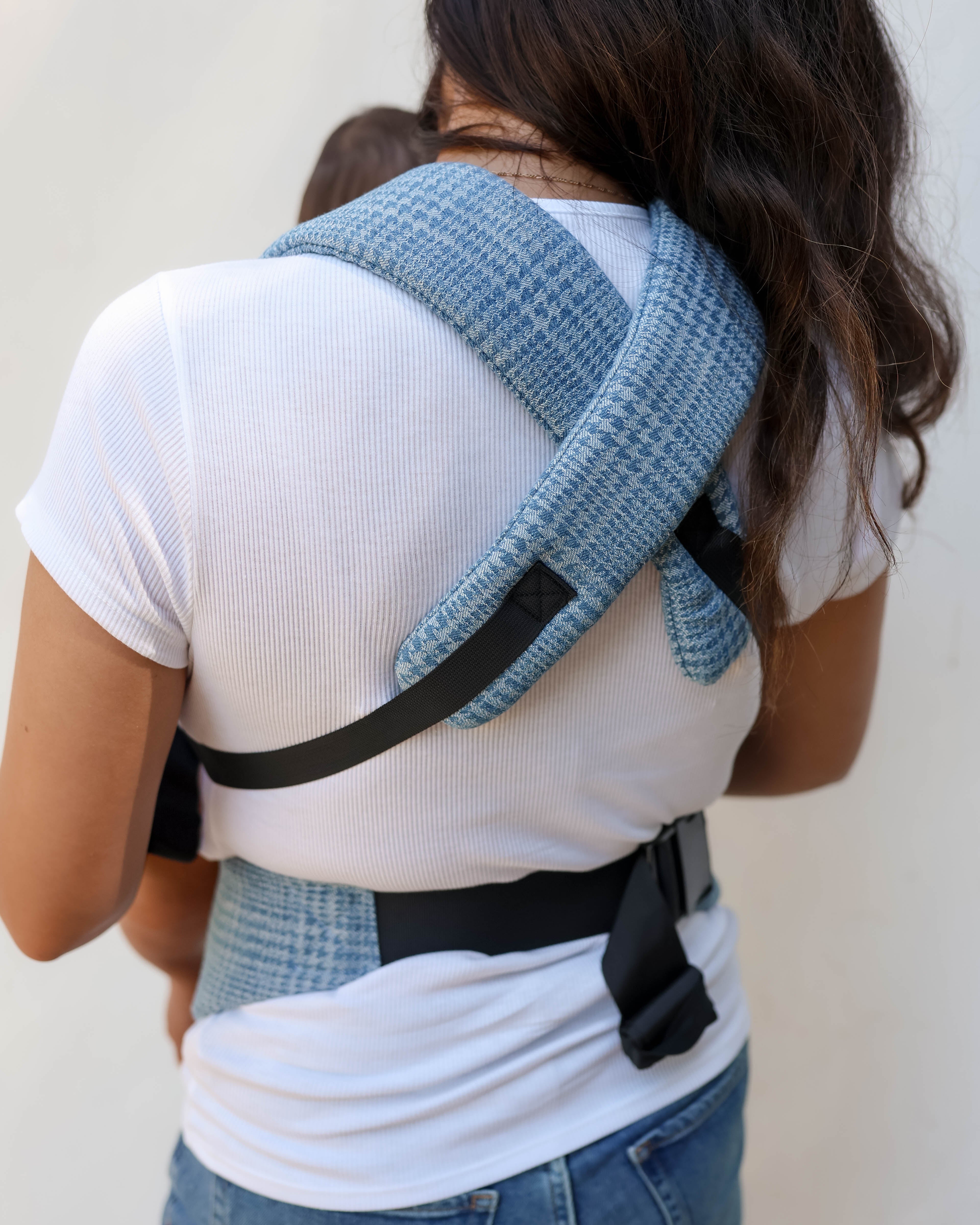 A woman shown from behind wearing a blue and white textured woven baby carrier wrap with black straps, holding a baby against her back.