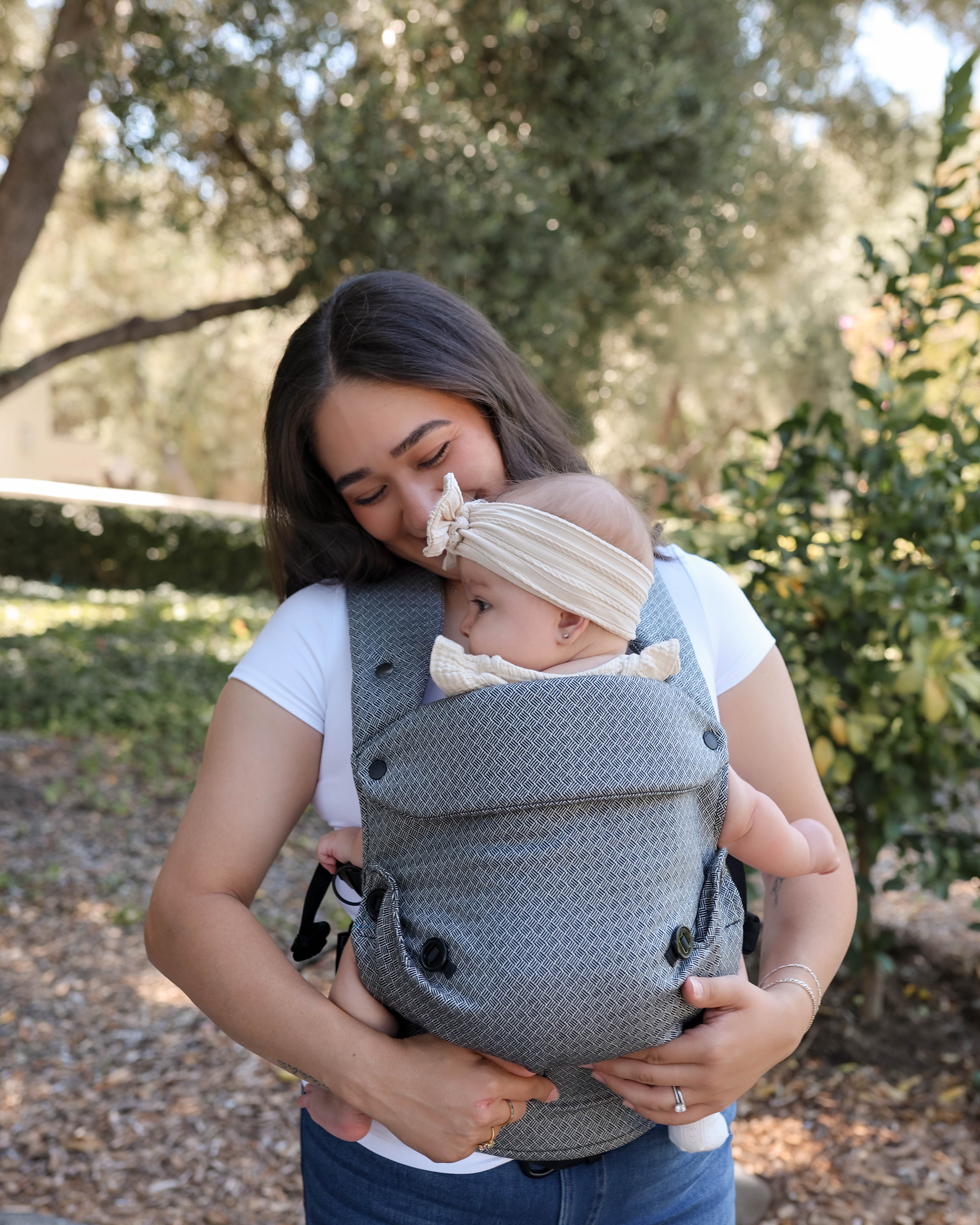 A woman outdoors wearing a black and white textured baby carrier with a baby inside, with a second baby wearing a cream headband resting on her shoulder.