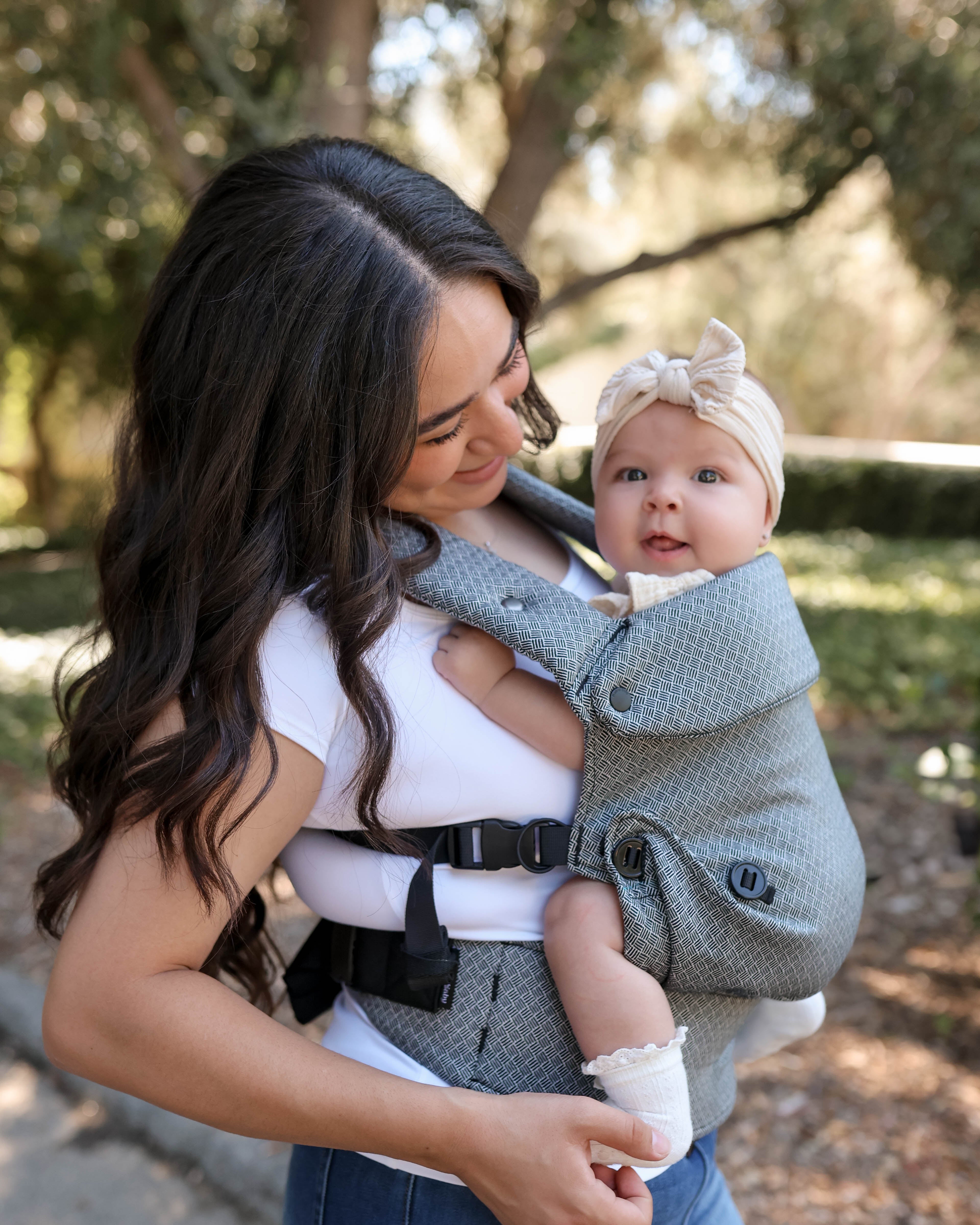 A woman outdoors wearing a baby carrier with a baby inside. The carrier is a textured black fabric with black straps and a black waist belt, and the baby is wearing a cream bow headband.