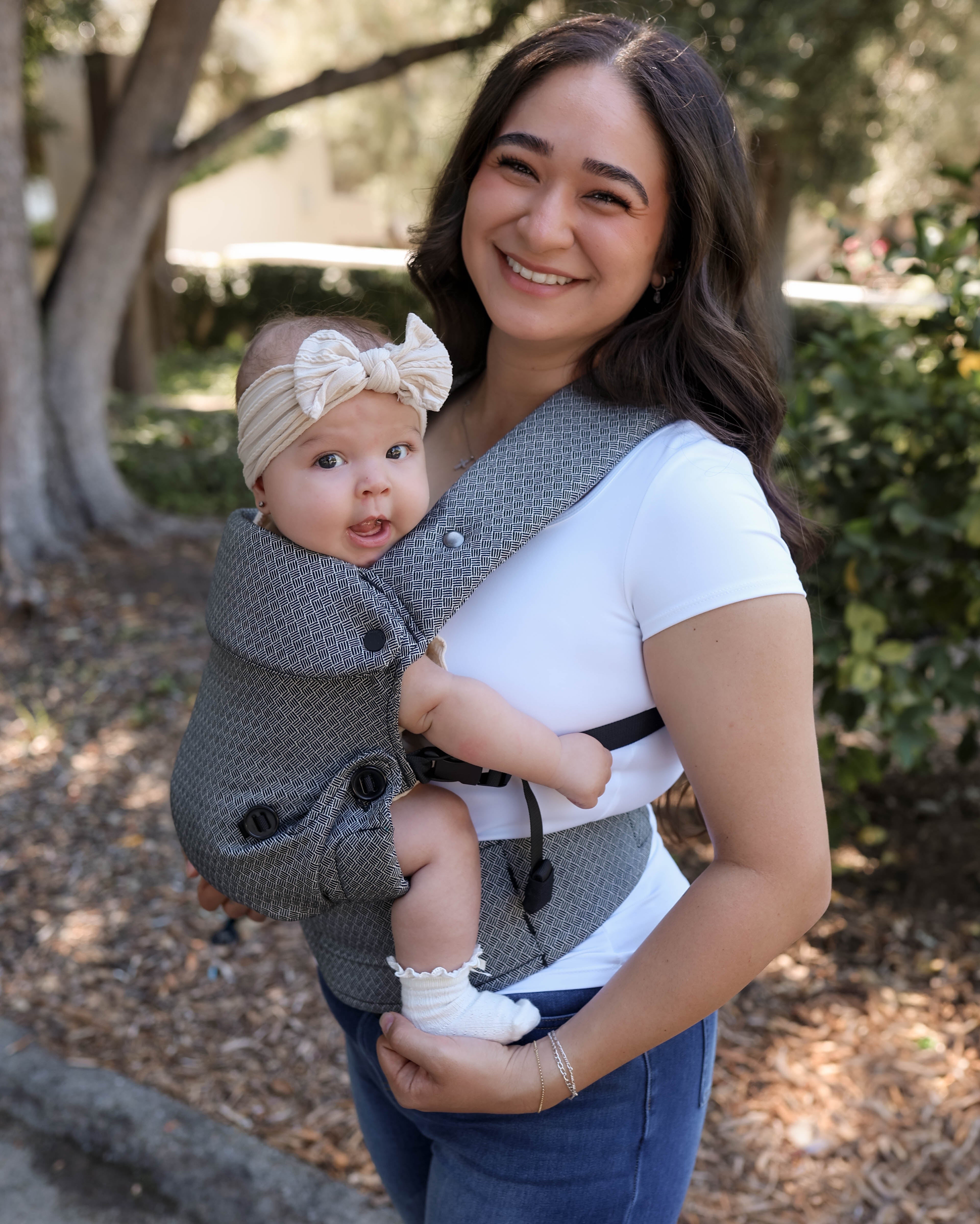 A woman outdoors wearing a black and white textured baby carrier with a baby inside. The baby is wearing a beige bow headband.