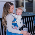 Woman holding a baby in a carrier and a coffee cup outdoors.