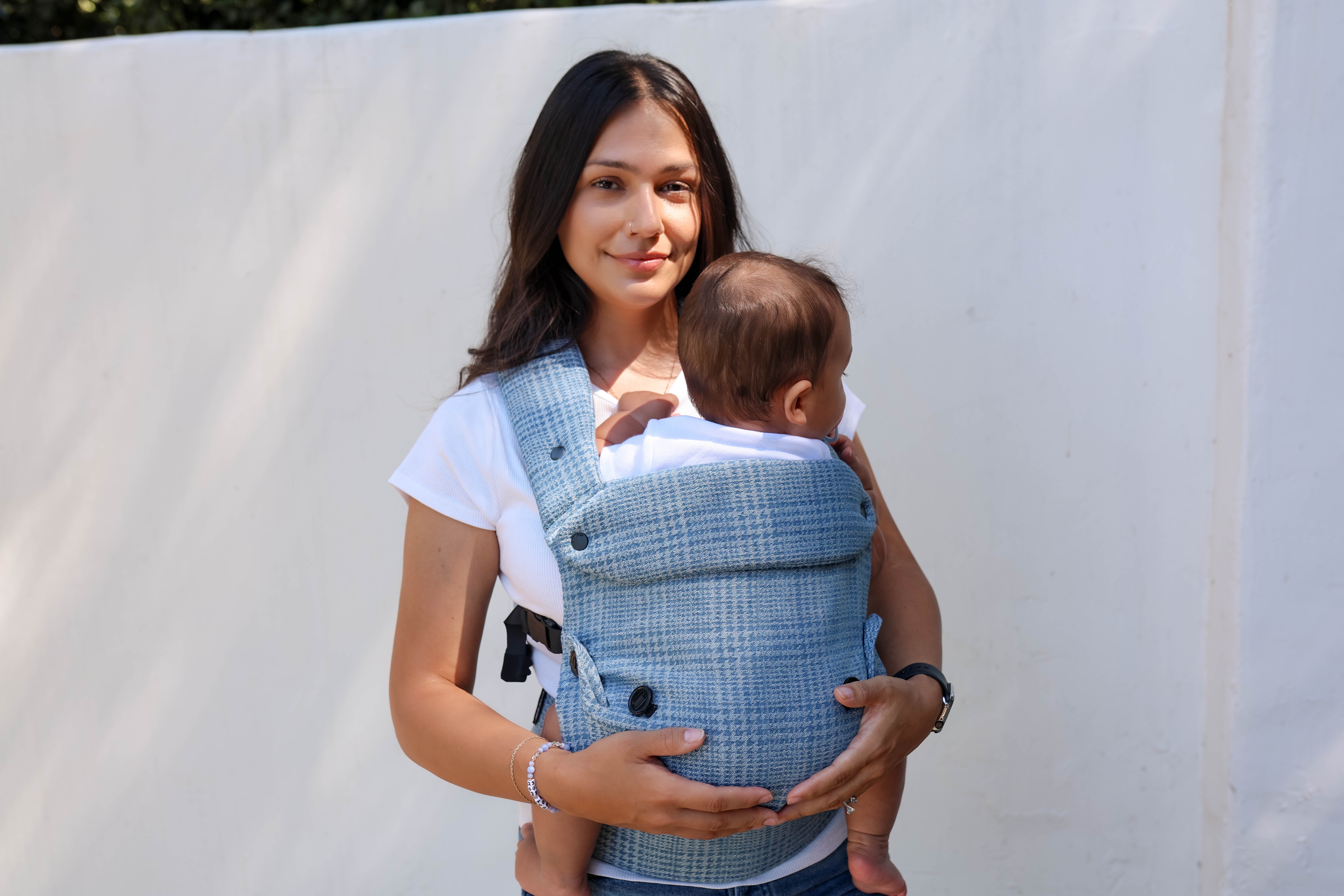A woman wearing a white t-shirt carries a baby in a blue and white plaid wrap-style baby carrier vest against a plain white background.