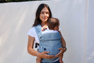 A woman wearing a white t-shirt carries a baby in a blue and white plaid wrap-style baby carrier vest against a plain white background.