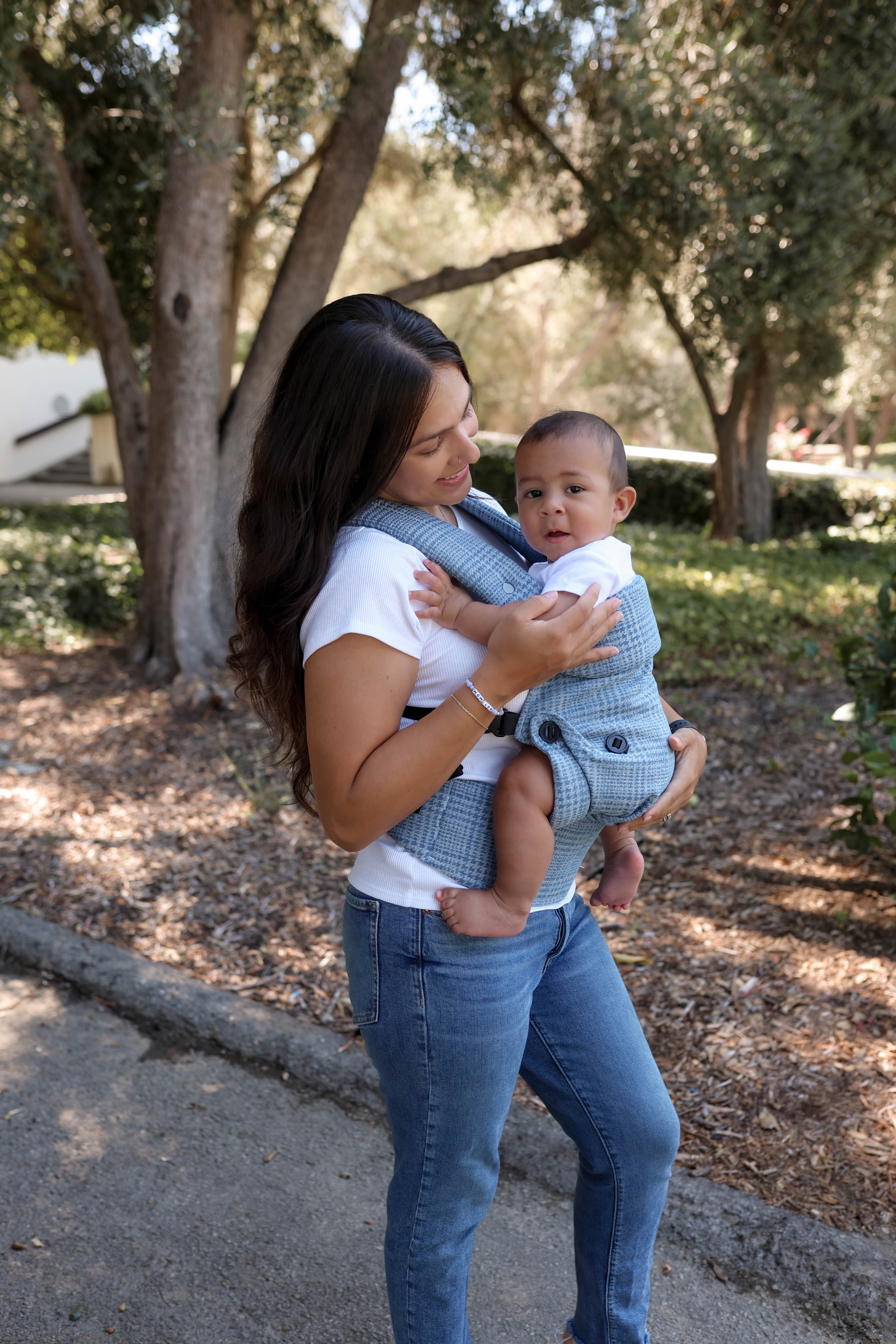 A woman outdoors carrying a baby in a blue and white checkered baby carrier wrap, set against a background of trees and a paved path.