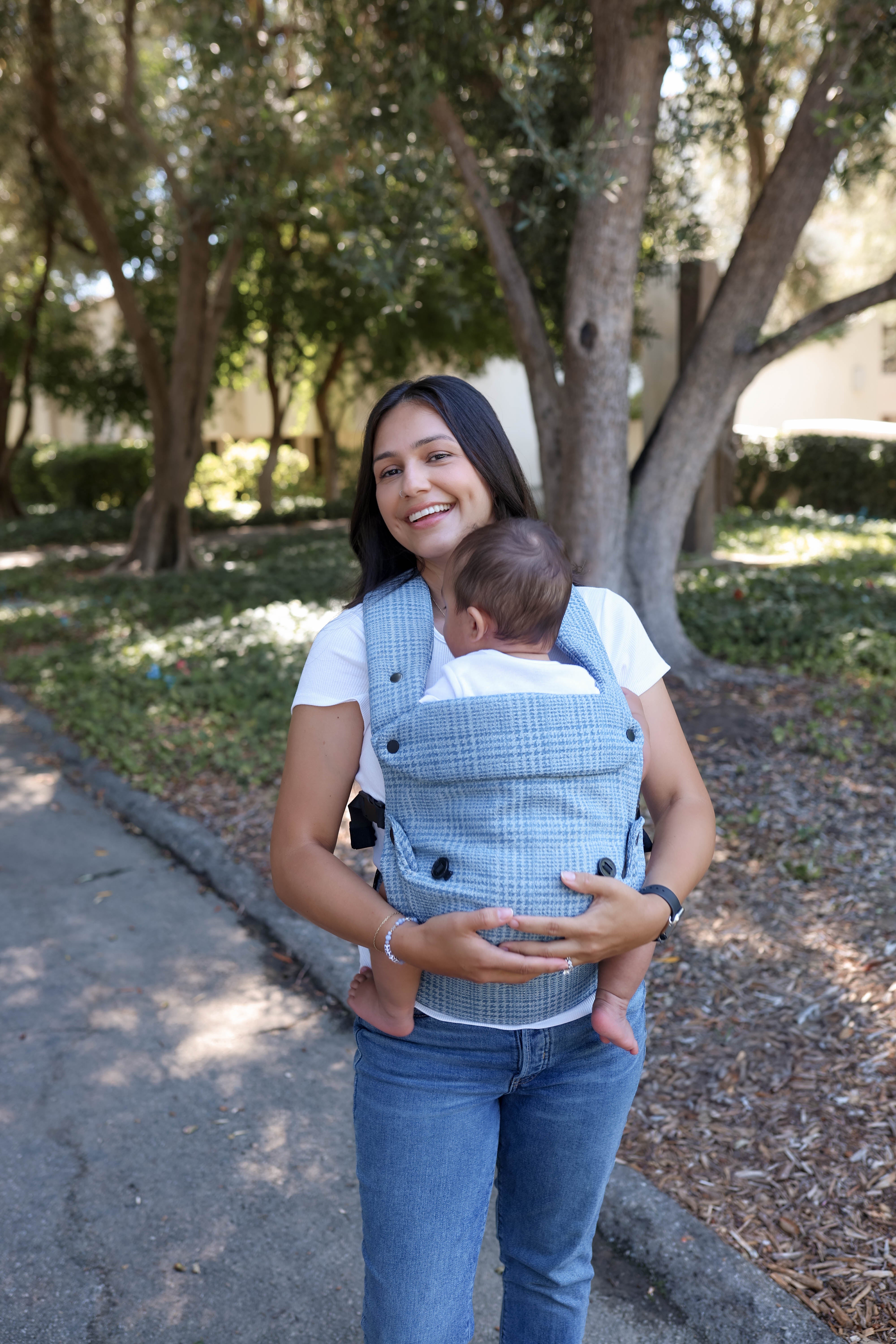 A woman wearing a light blue and white jacquard denim baby carrier with a baby inside, standing outdoors on a paved path with trees in the background.