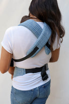 A woman shown from behind wearing a blue and white textured woven baby carrier wrap with black straps, holding a baby against her back.