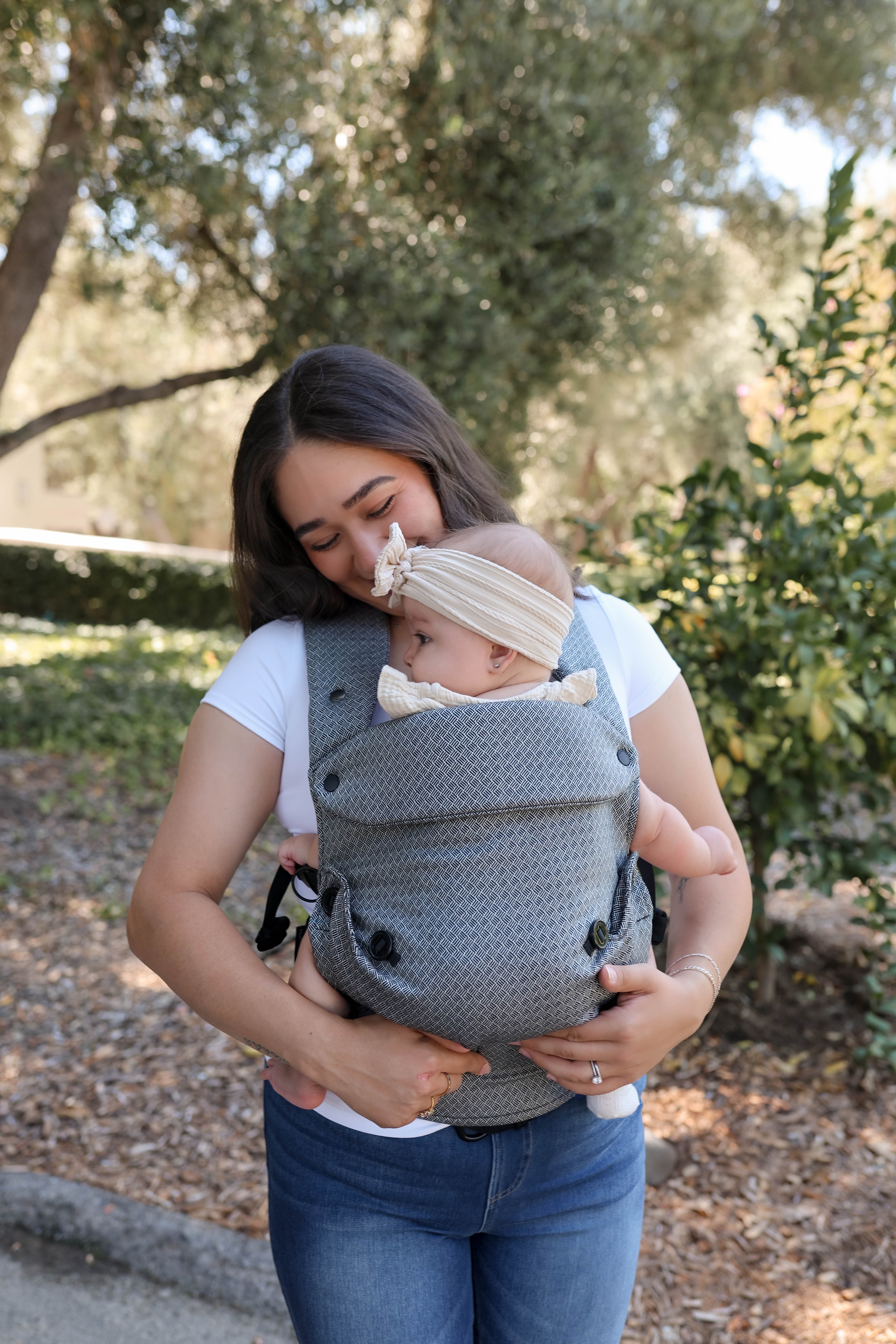 A woman outdoors wearing a black and white textured baby carrier with a baby inside, with a second baby wearing a cream headband resting on her shoulder.