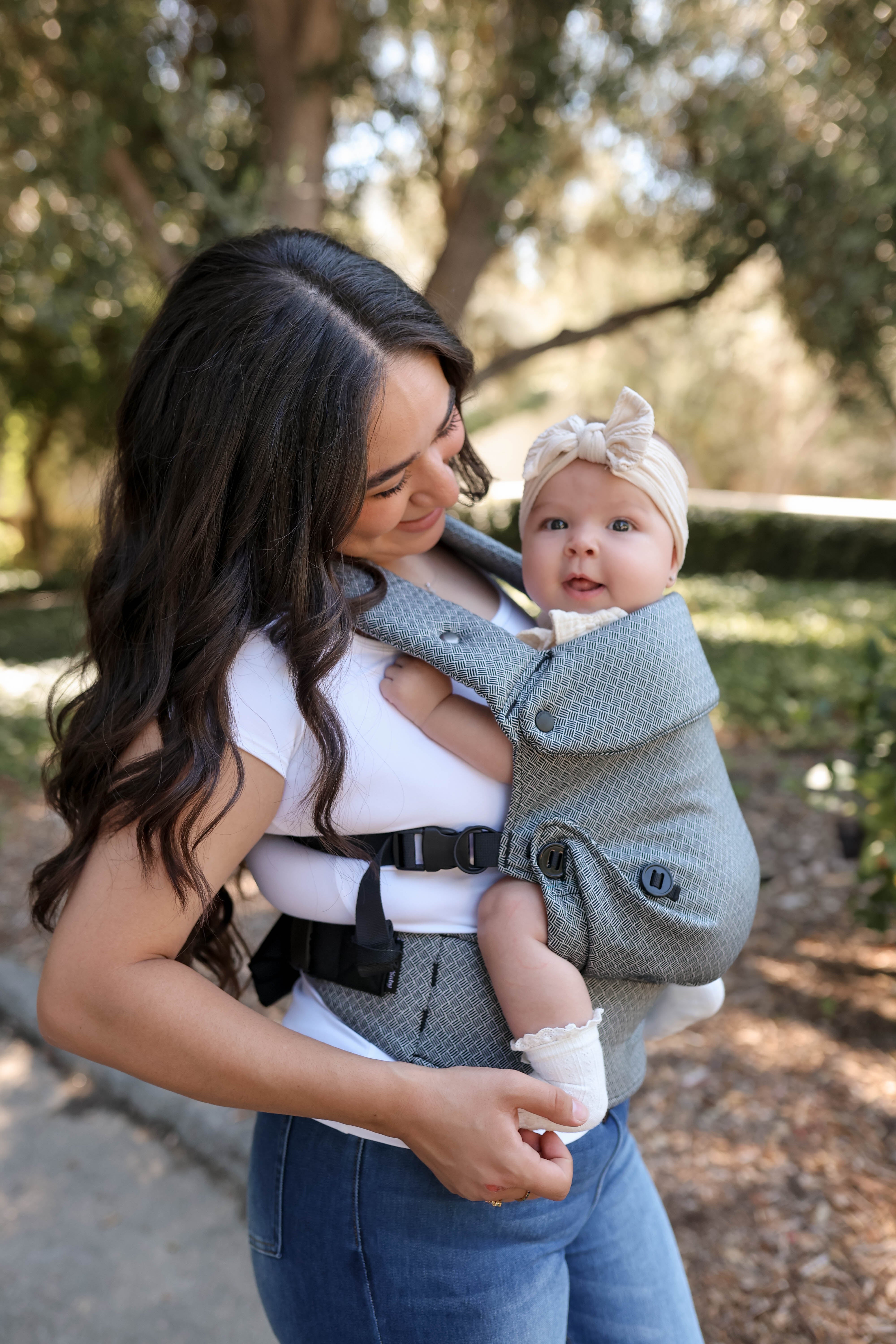A woman outdoors wearing a baby carrier with a baby inside. The carrier is a textured black fabric with black straps and a black waist belt, and the baby is wearing a cream bow headband.