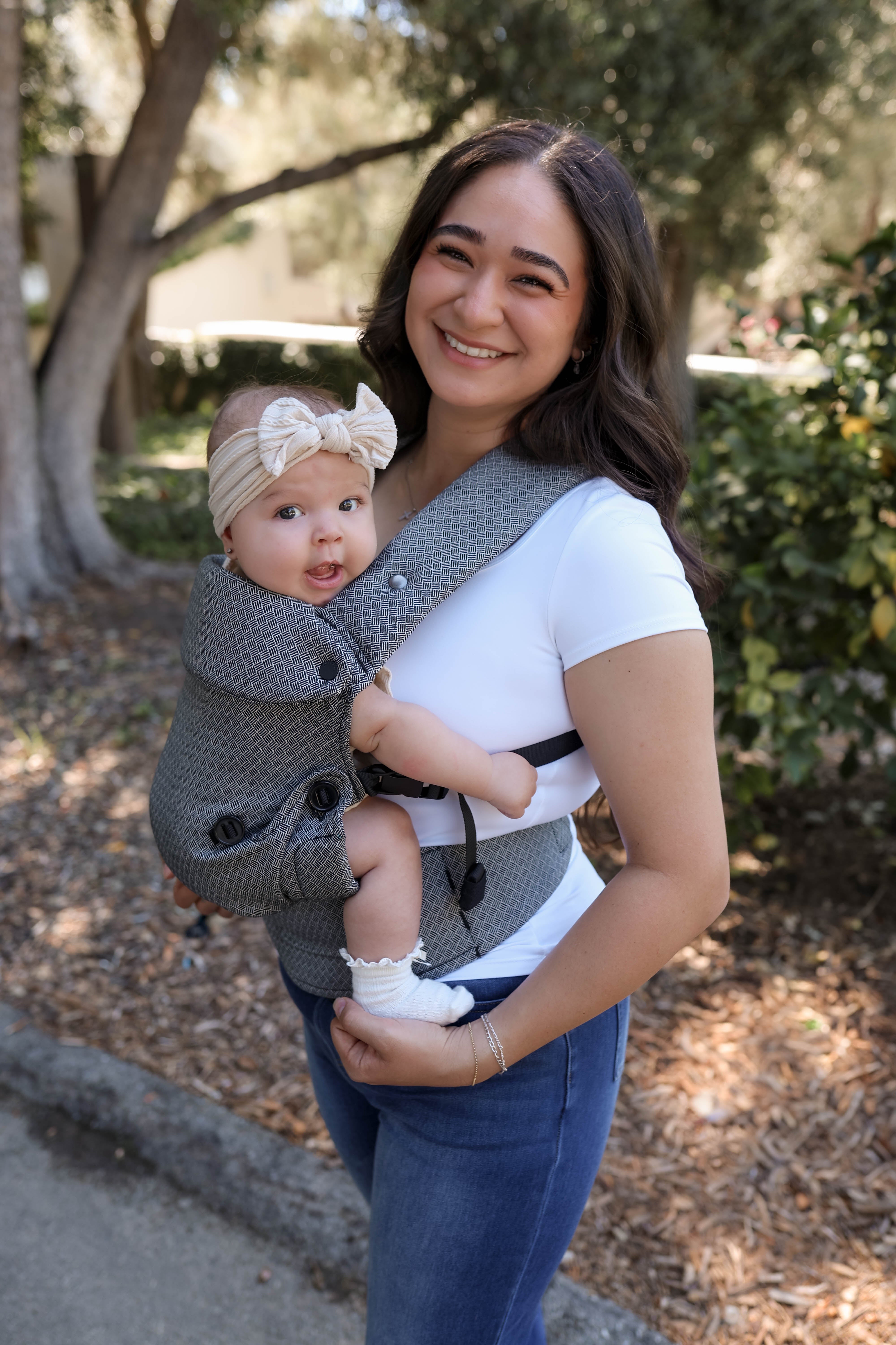 A woman outdoors wearing a black and white textured baby carrier with a baby inside. The baby is wearing a beige bow headband.